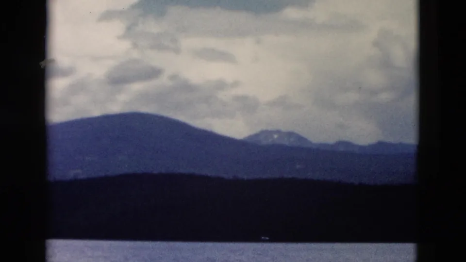 1958 Yellowstone 8mm frame showing a low mountain silhouette, dark forested shore, calm water, and soft clouds.