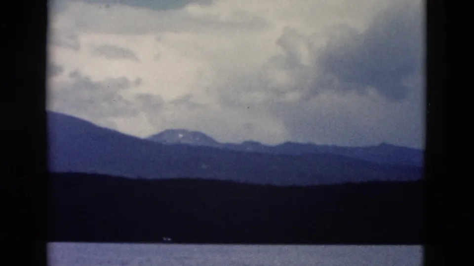 1958 Yellowstone home-movie frame with a distant ridge, snow patch, dark tree line, calm water, and overcast sky.