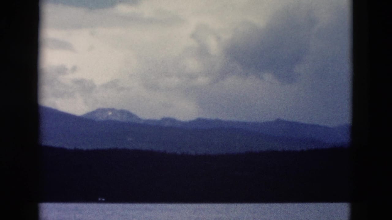 1958 Yellowstone home-movie frame showing distant mountains under heavy clouds with forest and water in the foreground.