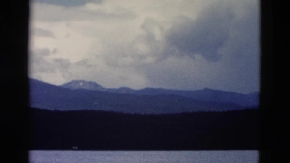 1958 Yellowstone home-movie frame showing distant mountains under heavy clouds with forest and water in the foreground.