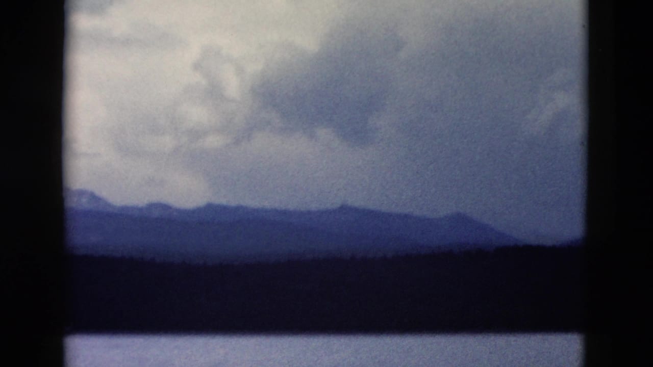 Yellowstone-area mountain view on 8mm film, with clouds above a dark forest and a calm strip of water.