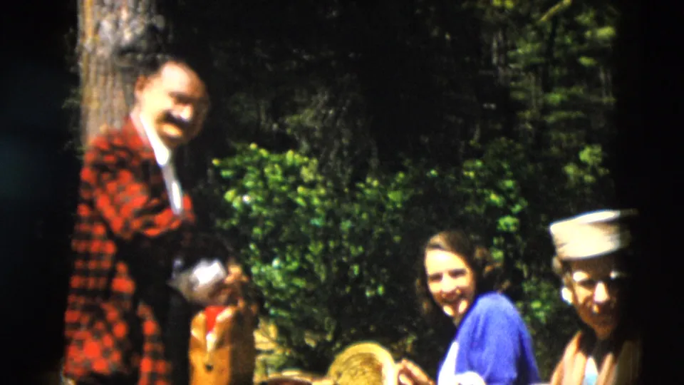 1958 Yellowstone home-movie frame of adults smiling at a picnic in the woods, with a red plaid jacket and sailor-style cap visible.