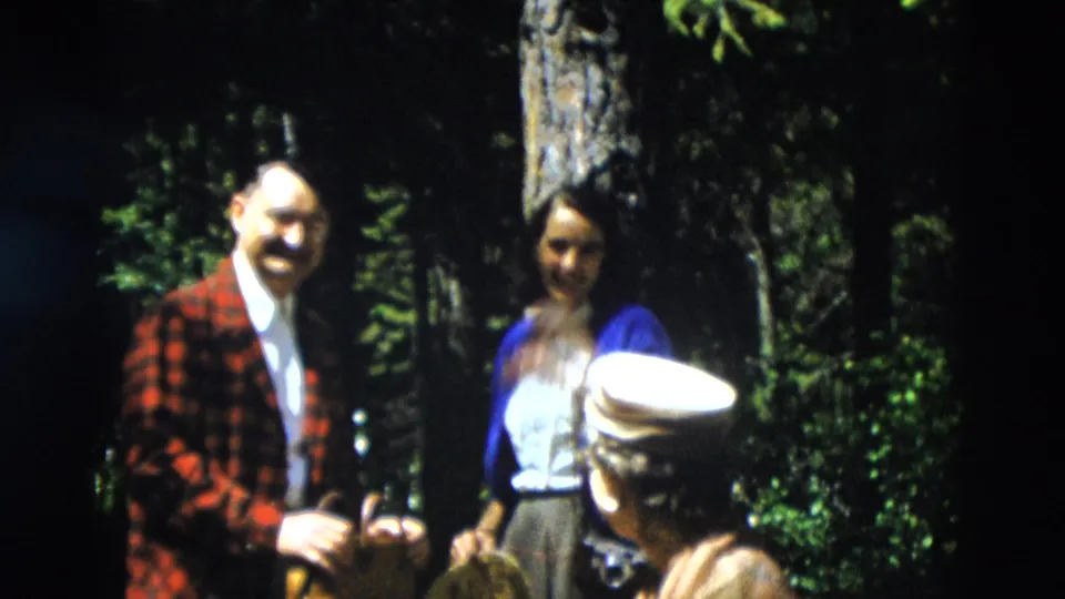 A man in a plaid jacket and a woman in a blue cardigan face the camera in the 1958 Yellowstone forest scene.