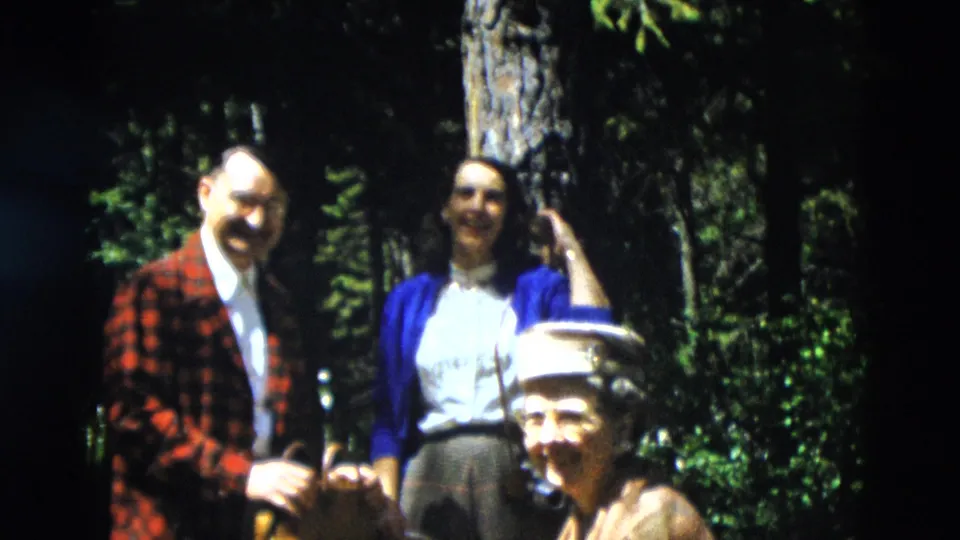 Three adults in 1958 Yellowstone footage gather near pine trees, with the seated woman smiling in the foreground.