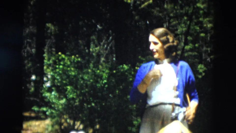 1958 Yellowstone home-movie frame showing a woman in a blue cardigan and skirt standing in a forested setting.