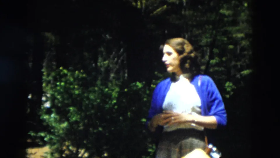 1958 Yellowstone frame showing a woman in a blue cardigan turned in profile, standing among shaded trees and foliage.