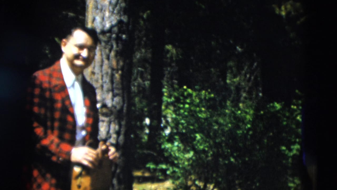Man in red plaid jacket standing beside a tree in a shaded forest.