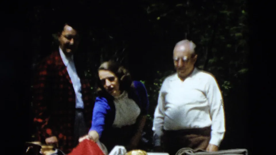 Three adults gather in a shaded Yellowstone forest, with a red plaid jacket, blue cardigan, white shirt, and cooler visible.