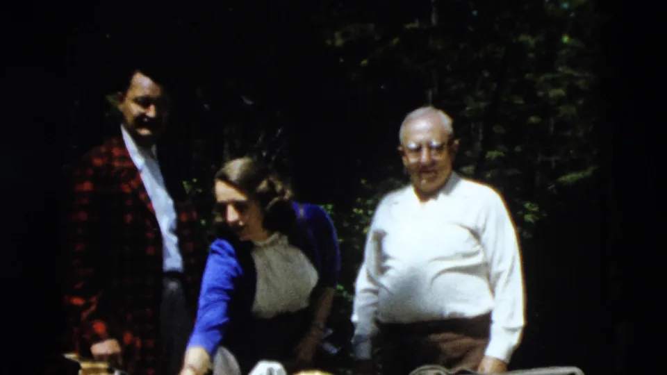 1958 8mm home-movie frame of two men and a woman standing by a table in a shaded wooded area during a travel stop.