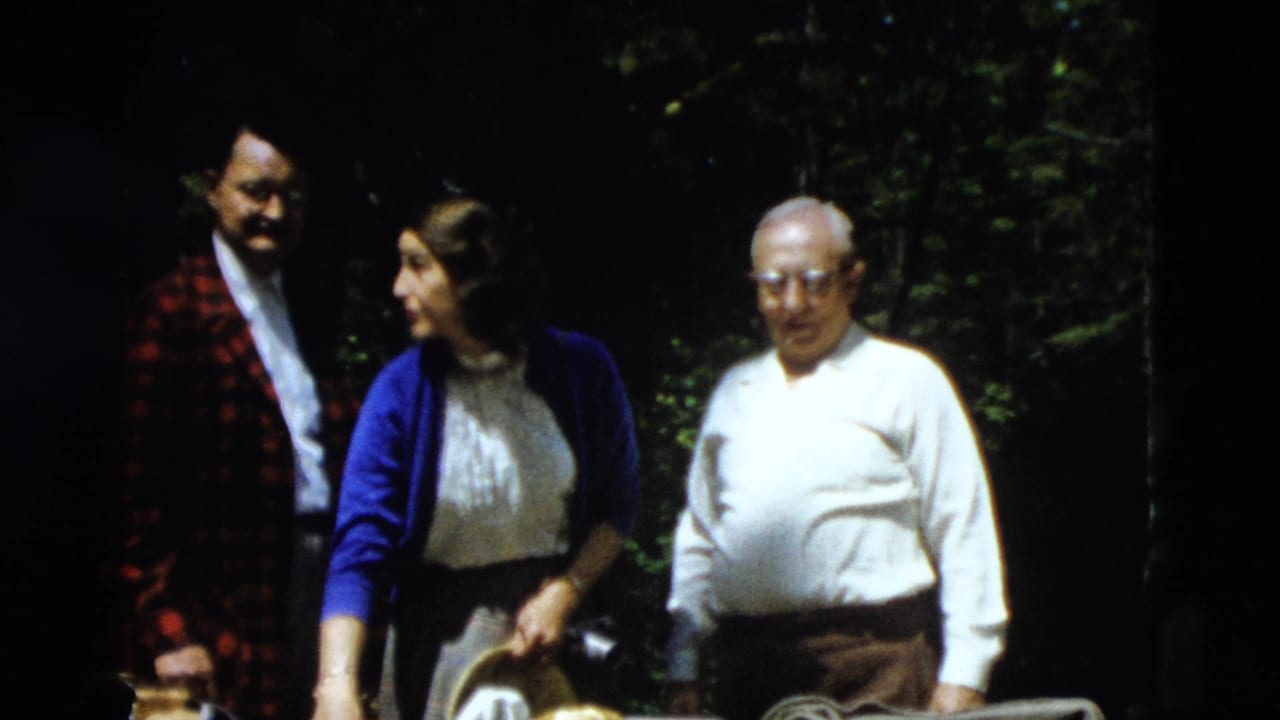 The woman in a blue cardigan looks toward the man in a red plaid jacket while an older man in a white shirt stands nearby.