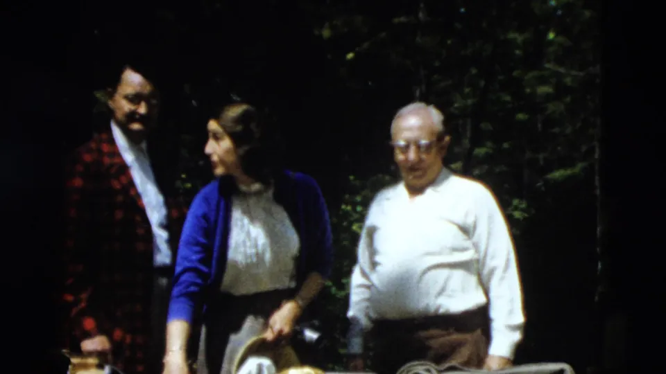 The woman in a blue cardigan looks toward the man in a red plaid jacket while an older man in a white shirt stands nearby.