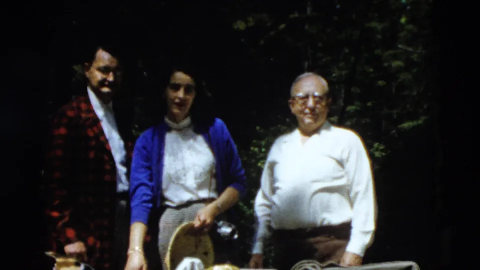 Three adults face the camera in a 1958 home-movie frame, with picnic items and dense trees behind them.