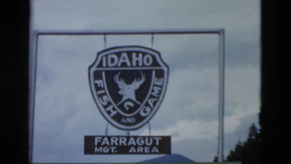 A roadside sign reads Idaho Fish and Game and Farragut Management Area, filmed in soft 8mm color with sky and trees behind it.