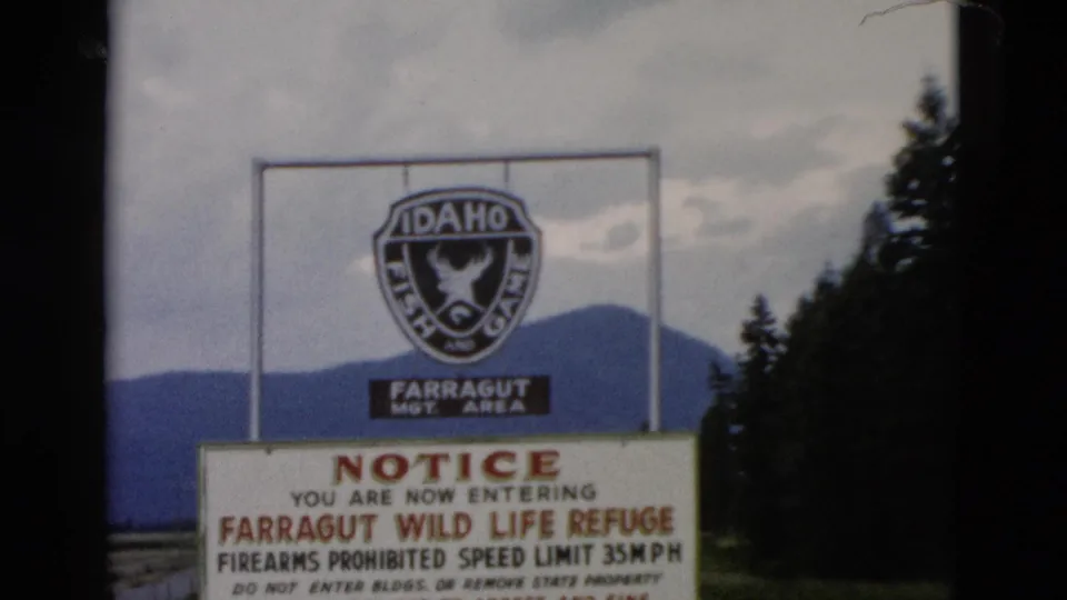 1958 8mm wider frame showing the Farragut management area sign, Idaho Fish and Game emblem, notice board, trees, and distant mountains.