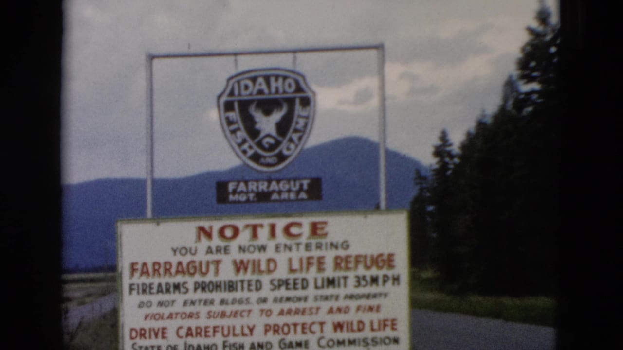 1958 8mm frame of a Farragut Wild Life Refuge entrance notice with an Idaho Fish and Game emblem, road edge, trees, and distant hills.