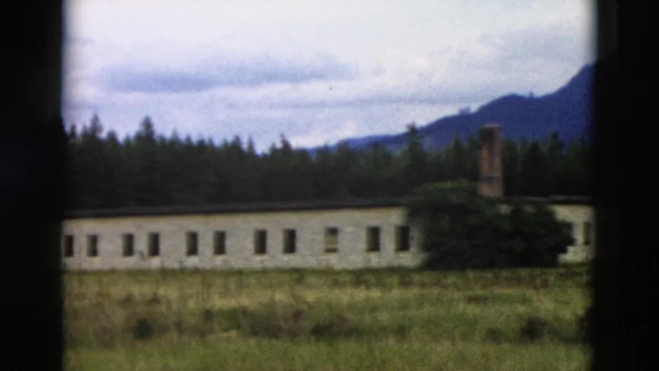 1958 Yellowstone 8mm still showing the right section of a long building with a brick chimney, shrubs, forest, and mountain slope.