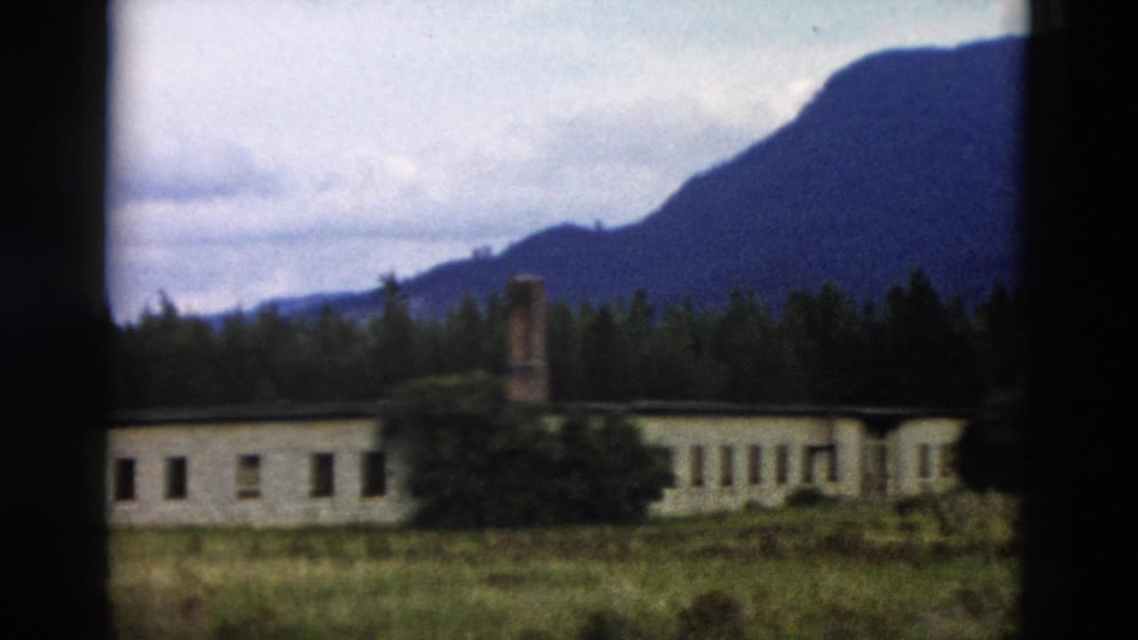 1958 Yellowstone-area 8mm frame showing a distant low building, brick chimney, grassy field, trees, and mountain backdrop.