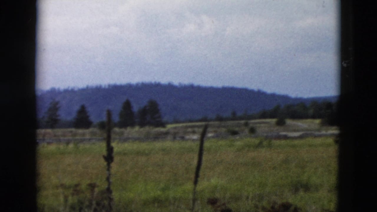 Window-framed field with fence posts, trees, and low hills under cloudy sky.