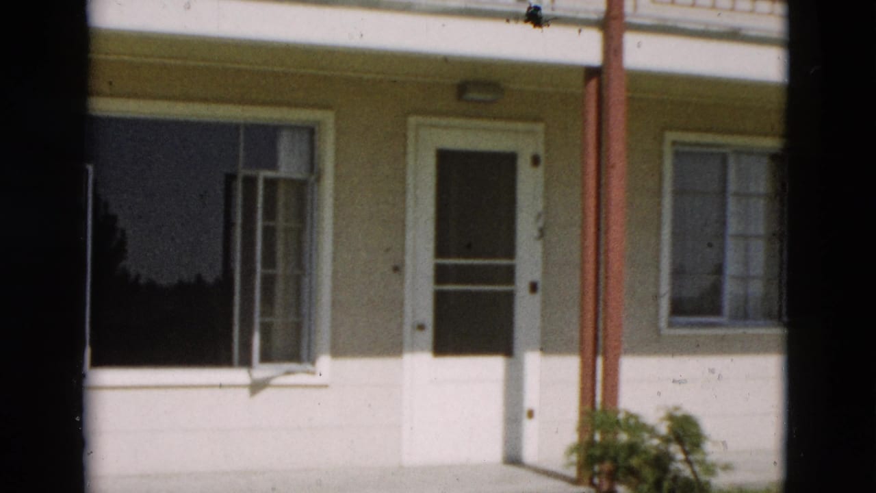1958 Yellowstone, Wyoming home-movie frame showing a pale house facade, white front door, open window, red porch posts, and a green plant.