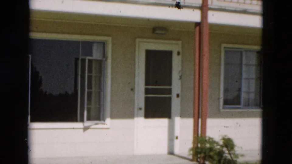 1958 Yellowstone, Wyoming home-movie frame showing a pale house facade, white front door, open window, red porch posts, and a green plant.