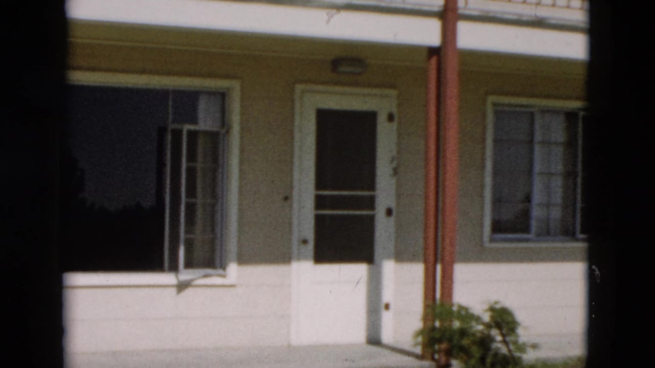 1958 Yellowstone, Wyoming frame of a modest house front with white door, open left window, curtained right window, red posts, and plant.