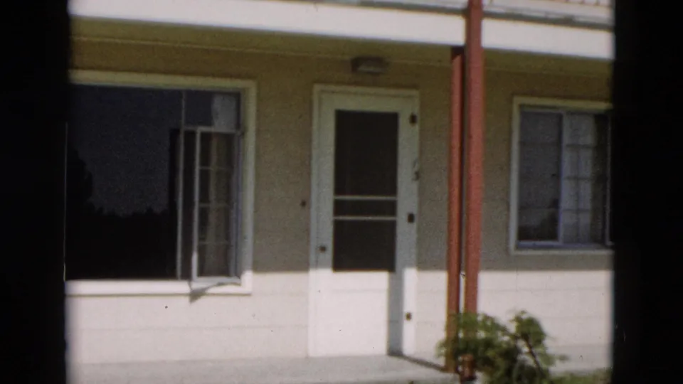 1958 Yellowstone, Wyoming frame showing a pale house exterior with white door, dark window reflections, red porch posts, and a small green plant.
