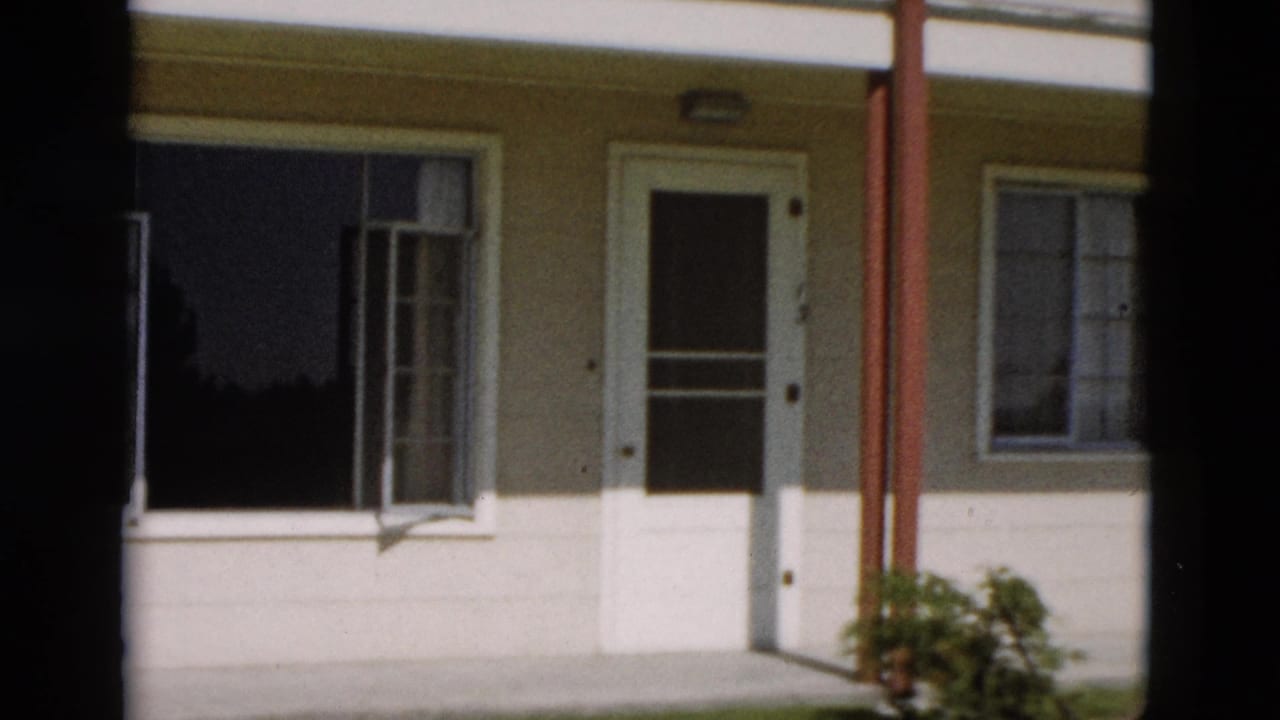 1958 8mm Yellowstone exterior showing a cream building side with a white door, open window, red support post, and small bush.