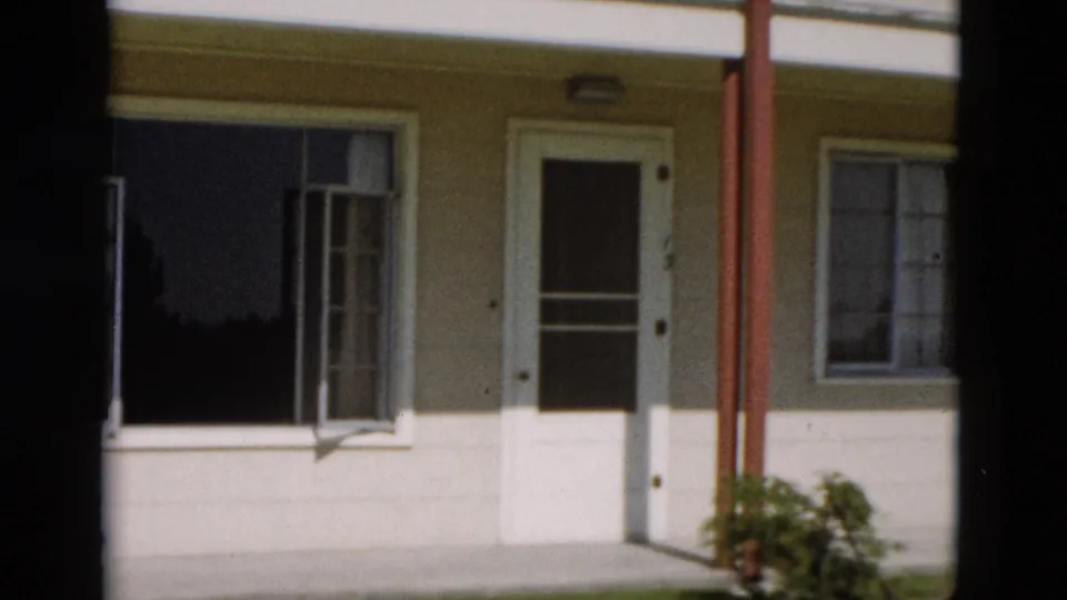1958 8mm Yellowstone exterior showing a cream building side with a white door, open window, red support post, and small bush.