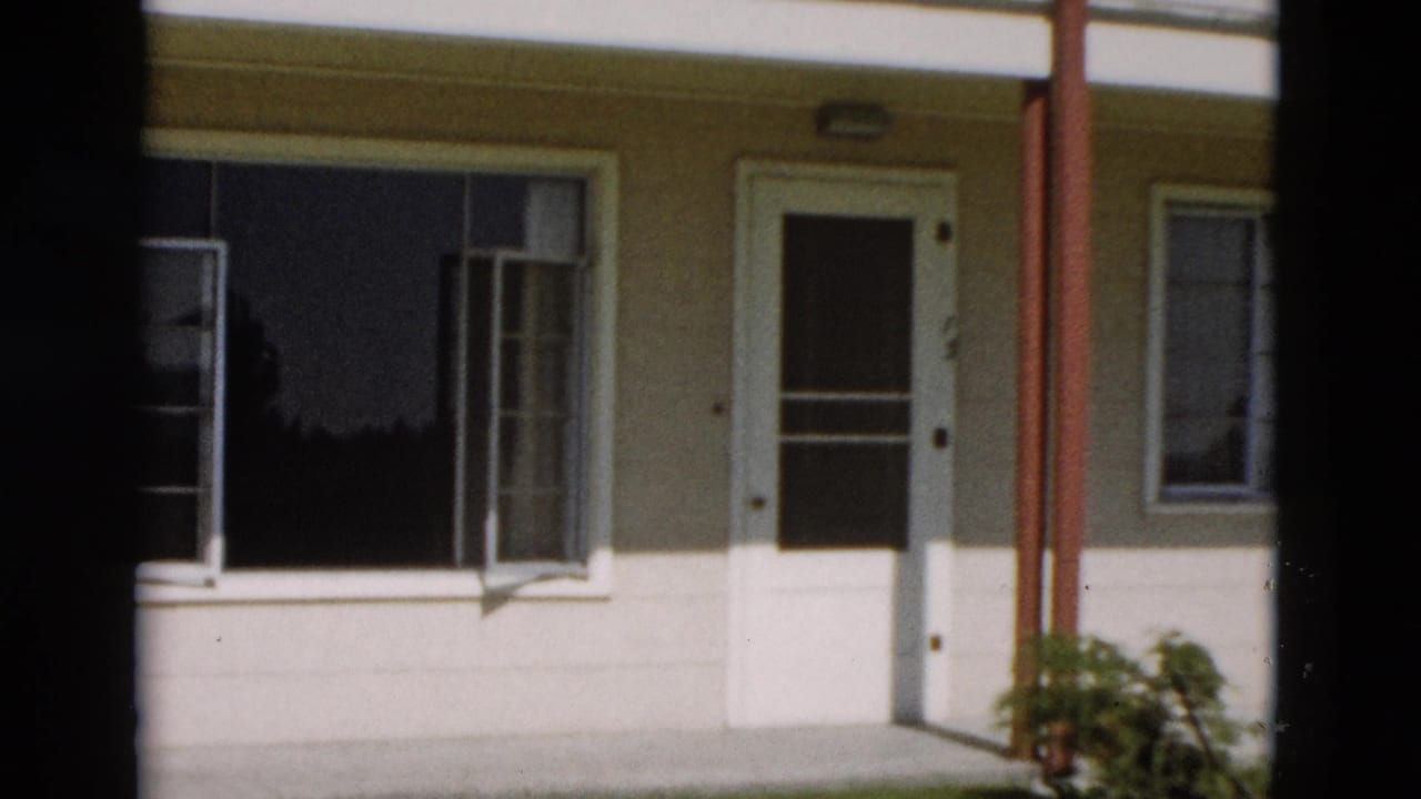 1958 Yellowstone home-movie frame of a cream exterior wall with white trim, open window, door, red posts, and bush.