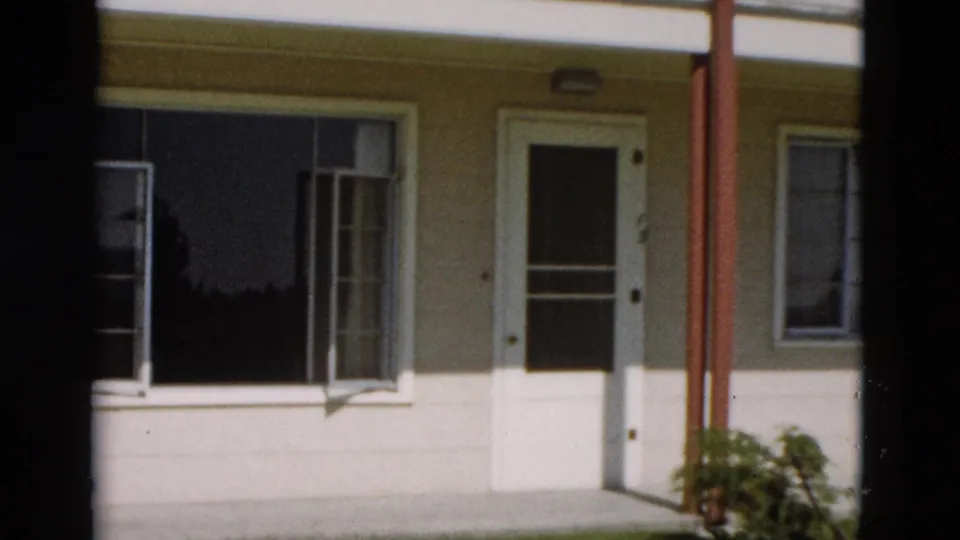 1958 Yellowstone home-movie frame of a cream exterior wall with white trim, open window, door, red posts, and bush.