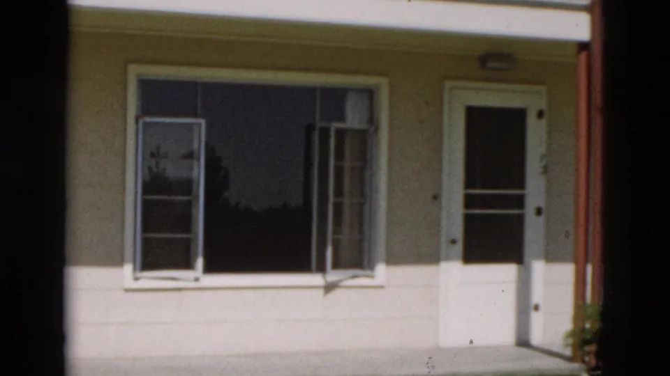 Final 1958 Yellowstone frame showing the cream building side with a large window, white door, red post, and reflected dark glass.