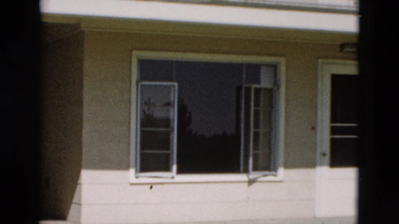 1958 Yellowstone home-movie frame showing open casement windows on a pale sunlit building wall with dark interior reflections.