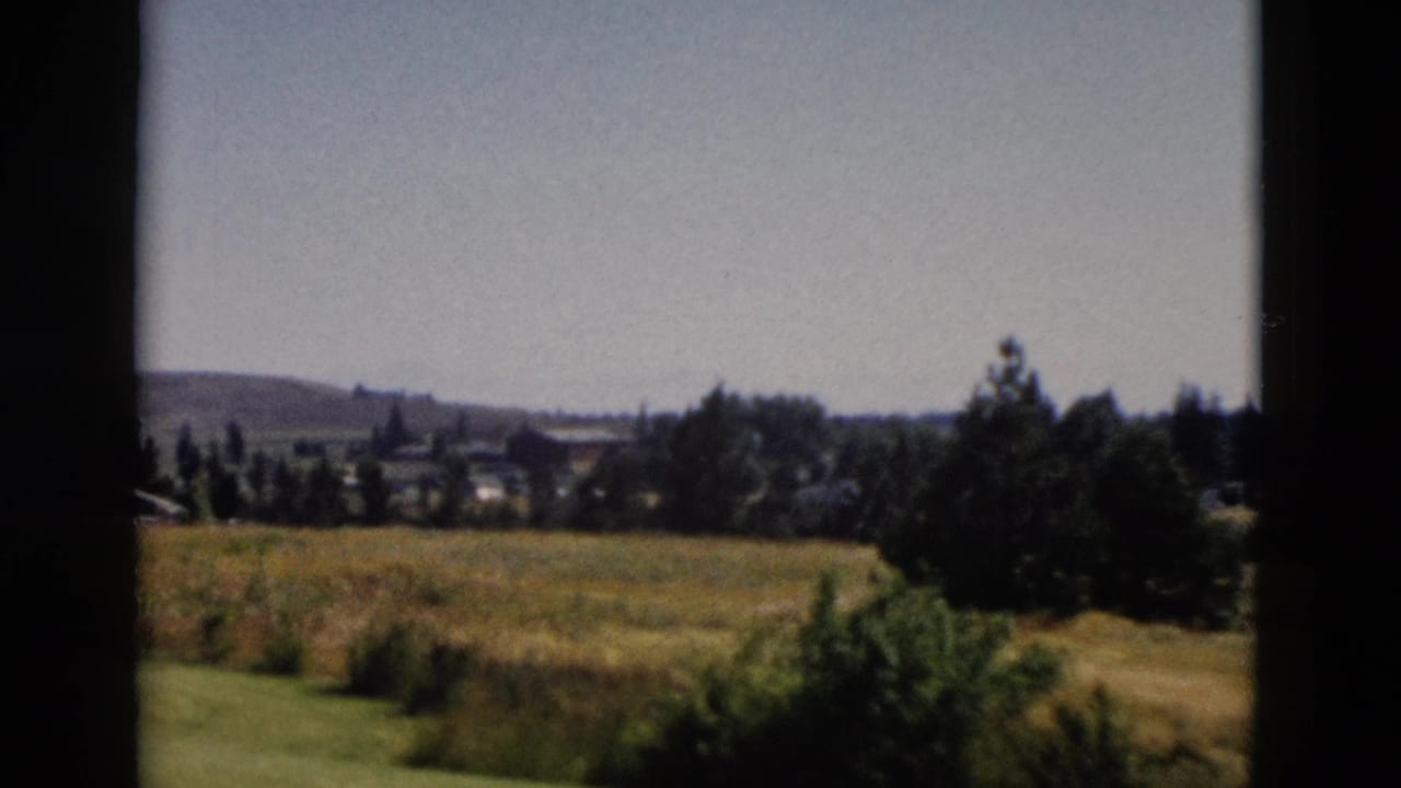 1958 Yellowstone-area landscape view with golden grass, shrubs, distant trees, small settlement shapes, and low hills.