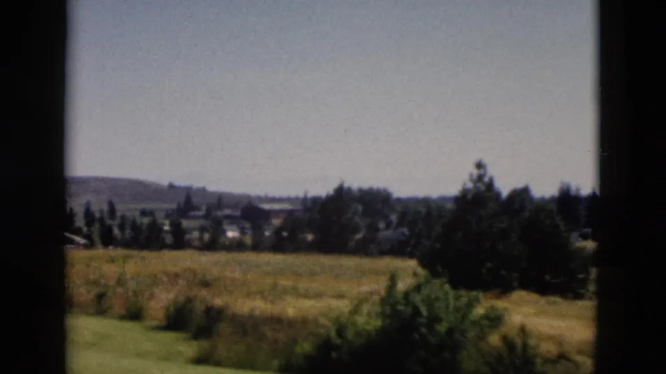 1958 Yellowstone-area landscape view with golden grass, shrubs, distant trees, small settlement shapes, and low hills.