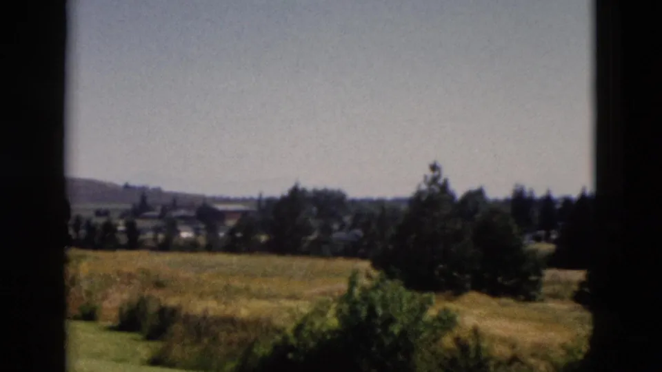 1958 Yellowstone-area 8mm frame showing open grassland, a distant settlement, treeline, low hill, and pale sky.