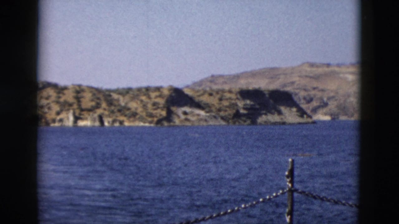 1958 8mm Yellowstone-area view across blue water toward rocky hills, framed by dark film borders and a chain railing.