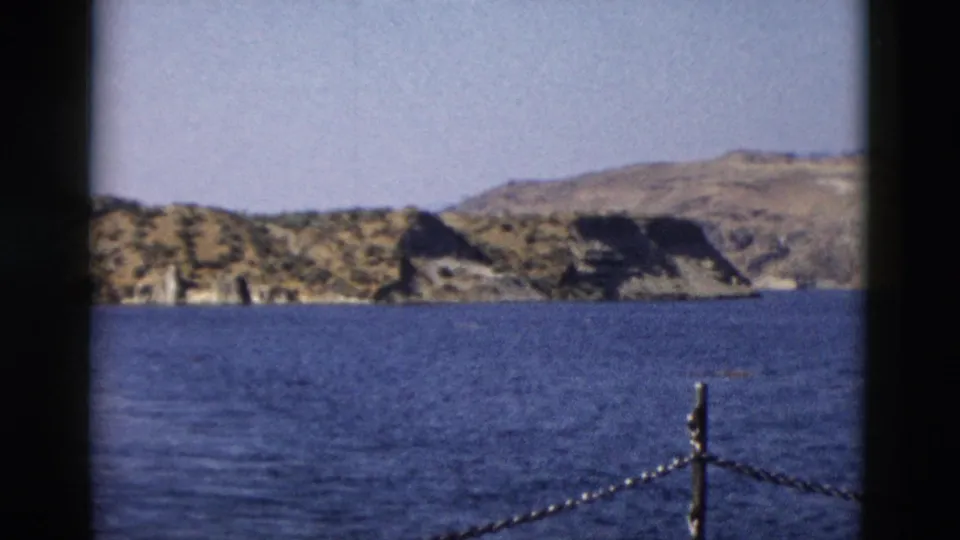 1958 8mm Yellowstone-area view across blue water toward rocky hills, framed by dark film borders and a chain railing.