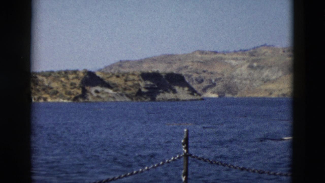 Rocky shoreline across blue water with a chain post and dark film borders.
