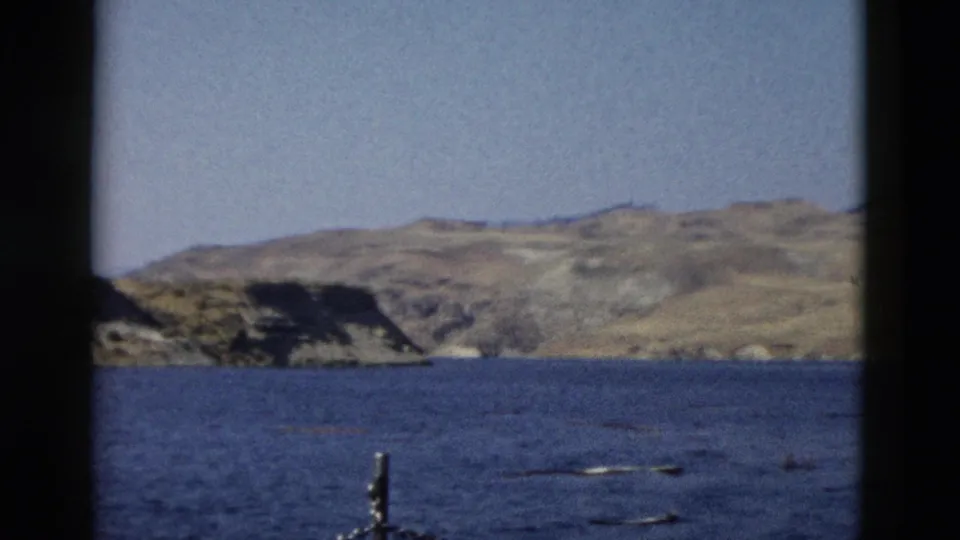 1958 home-movie view of blue water and rugged Wyoming hills, with soft grain and a chain railing at the lower edge.