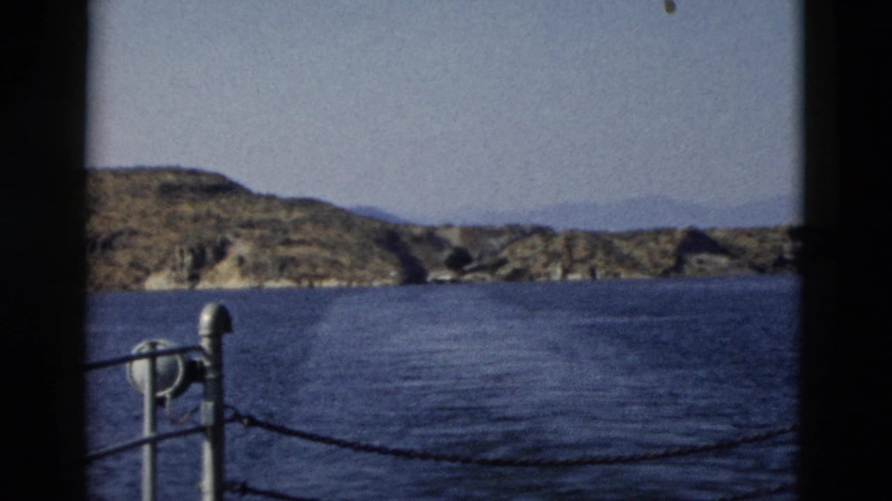 1958 8mm boat-view frame with blue water, rocky shoreline, hazy hills, metal post, and chain railing in the foreground.