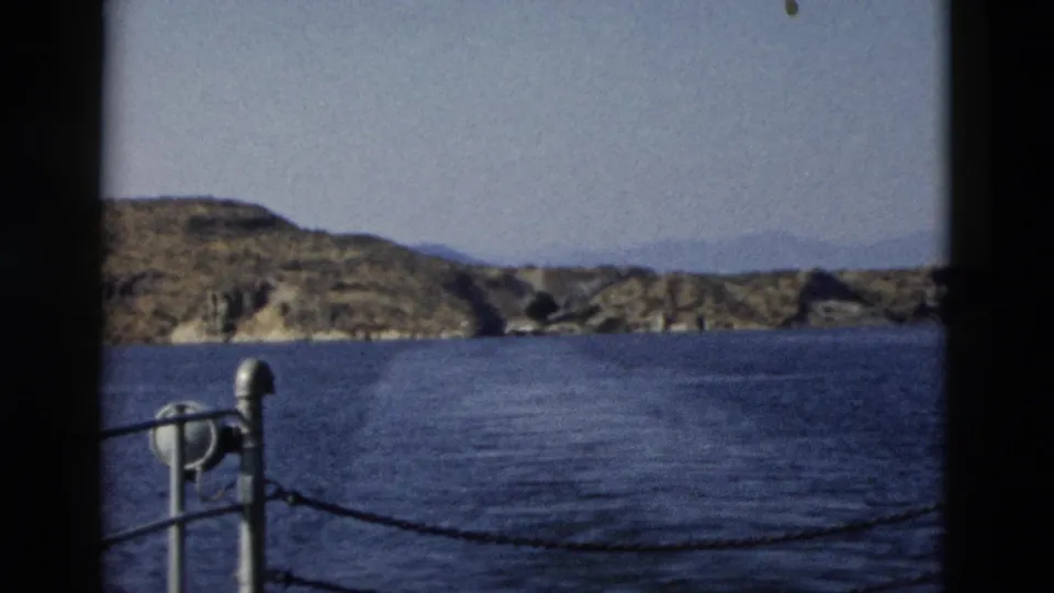 1958 8mm boat-view frame with blue water, rocky shoreline, hazy hills, metal post, and chain railing in the foreground.
