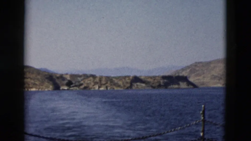 1958 8mm boat-view frame with open blue water, tan rocky shoreline, distant hills, a chain railing, and dark film-window borders.