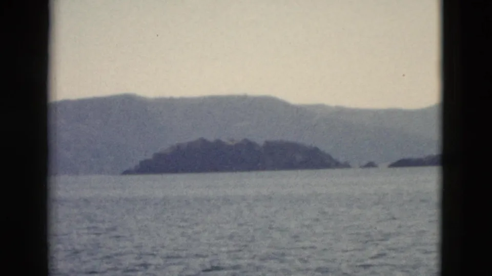 1958 8mm frame looking over calm water toward a dark island-like landform and hazy mountain ridges under a pale sky.