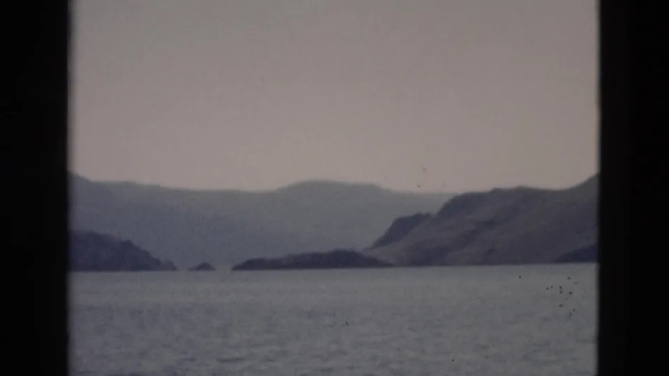 1958 Yellowstone-area lake view with distant mountain ridges, calm water, gray sky, and dark 8mm frame edges.