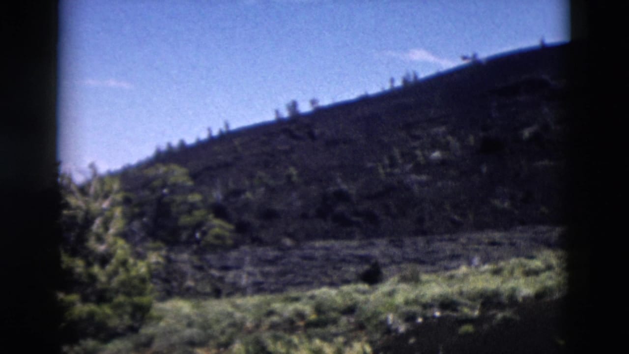 1958 Yellowstone home-movie frame showing a rugged dark hillside, blue sky, and sparse roadside vegetation.