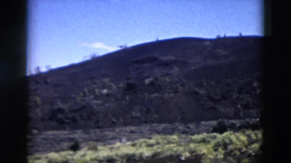 1958 Yellowstone view of a rolling rocky hill with scattered shrubs and a bright blue sky above.