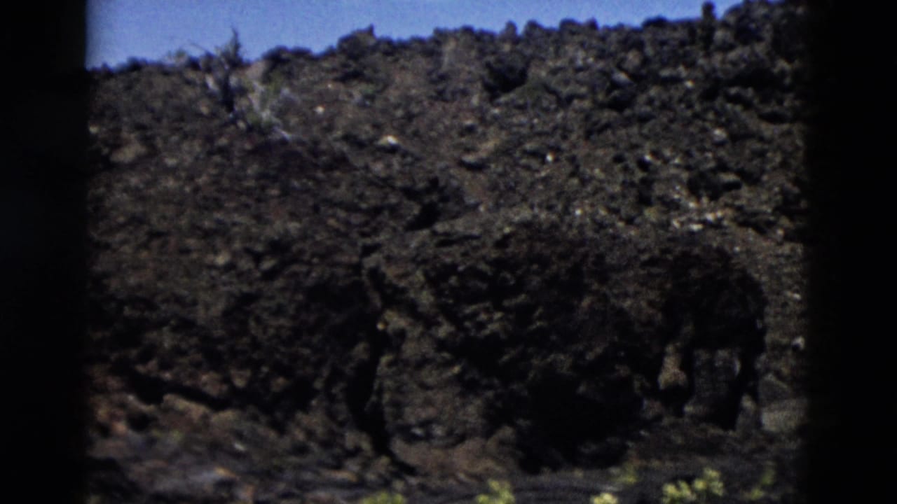 Rocky hillside with sparse scrub under blue sky, framed by dark film edges.