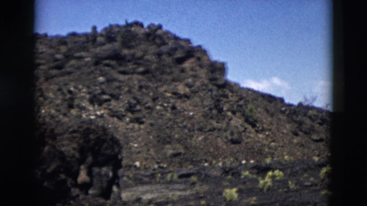 1958 Yellowstone home-movie frame showing a dark volcanic slope, scattered low plants, and blue sky.
