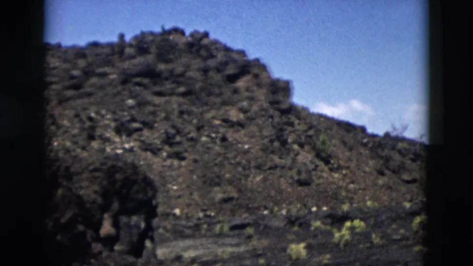 1958 Yellowstone home-movie frame showing a dark volcanic slope, scattered low plants, and blue sky.