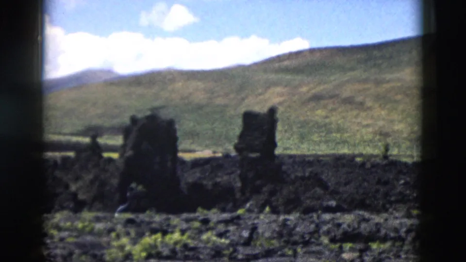 1958 Yellowstone home-movie view of dark volcanic rocks across rough ground with green hills beyond.